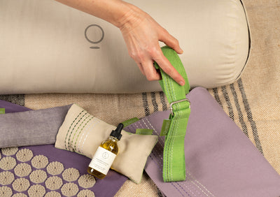 A hand holding green yoga strap over a beige bolster, body oil and an eye pillow.